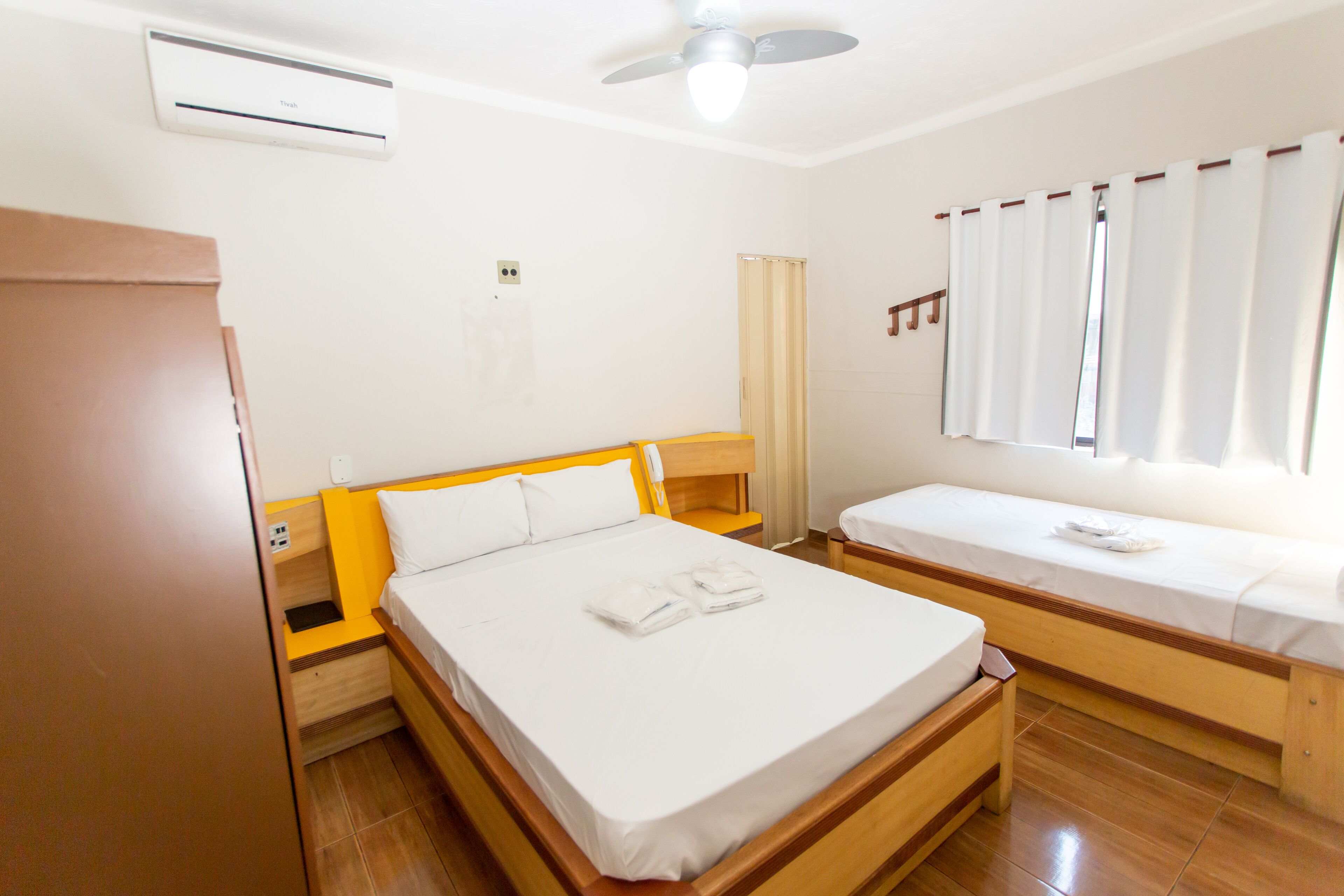 Habitación de hotel con una cama grande, una cama más pequeña, un armario, una mesita de noche, un equipo de aire acondicionado y una ventana con cortinas.