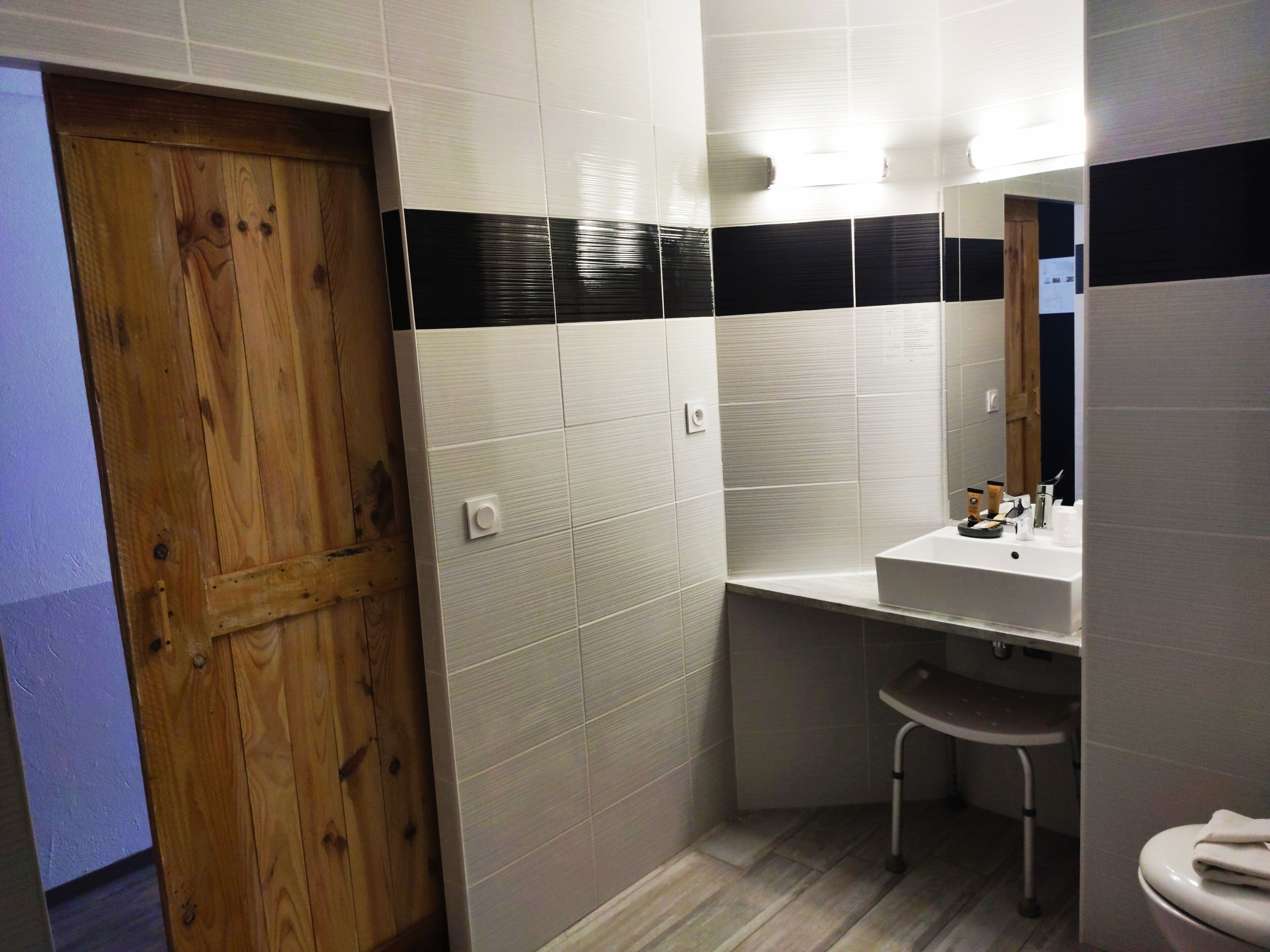 A bathroom with a wooden door, white and black tiled walls, a white sink, and a toilet.