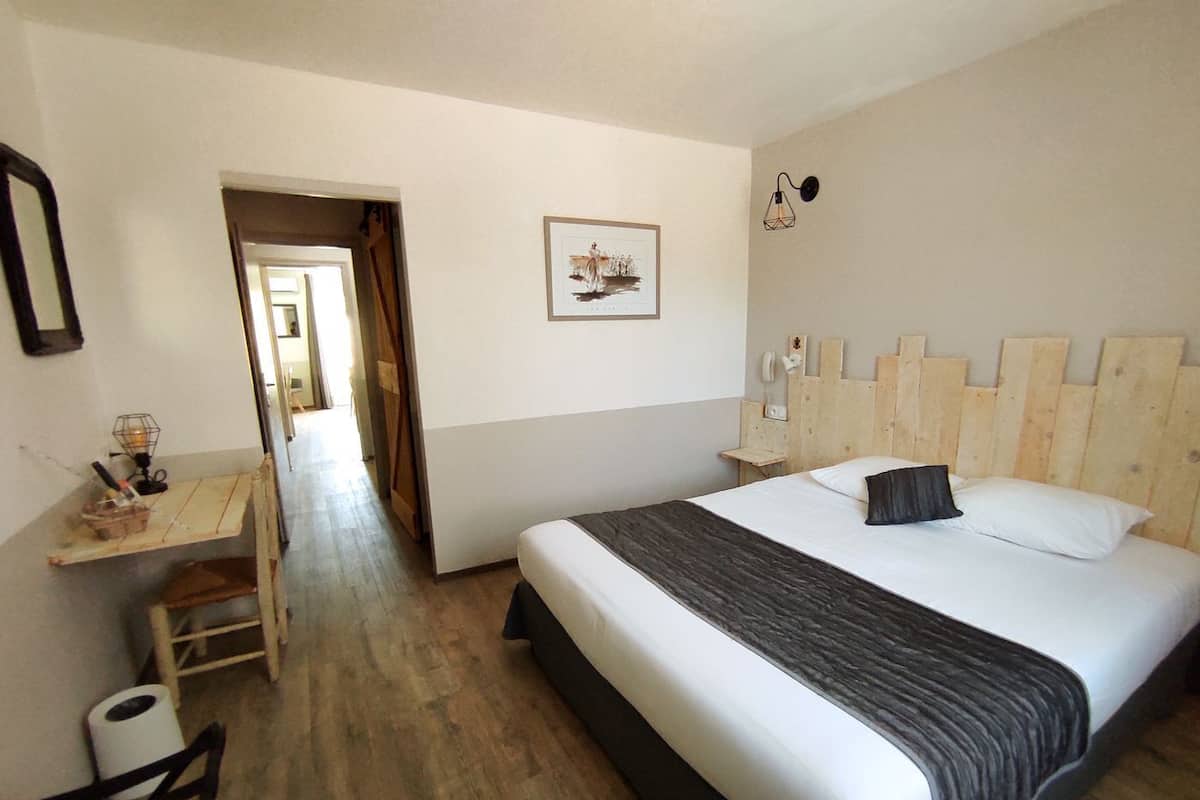 A bedroom with a wooden headboard, a bed with white bedding and a dark throw, a desk with a chair, and a framed picture on the wall.