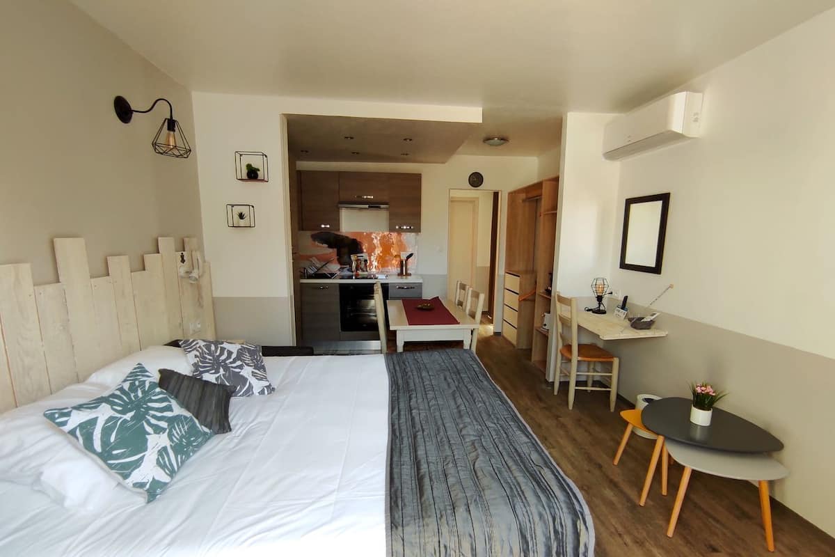 A hotel room with a bed, a desk, a chair, and a small table. There is a kitchenette in the background.