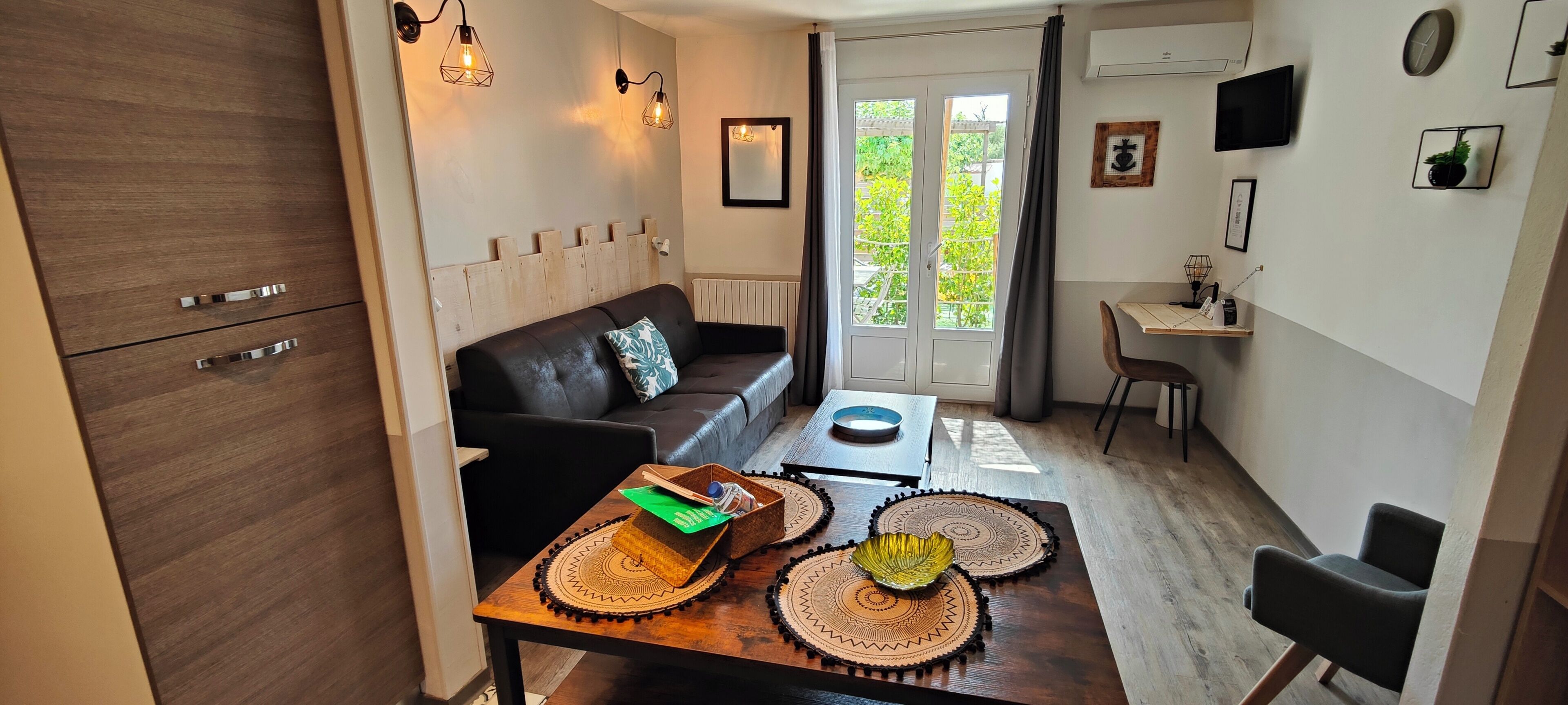 A modern living room with a sofa, a dining table with placemats, a small kitchen area, and a view through a glass door.