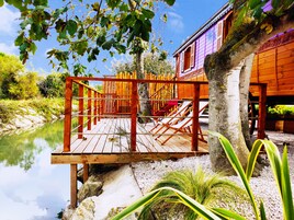 Una terraza de madera con vista a un río, un edificio de madera con contraventanas rojas y un árbol en primer plano.