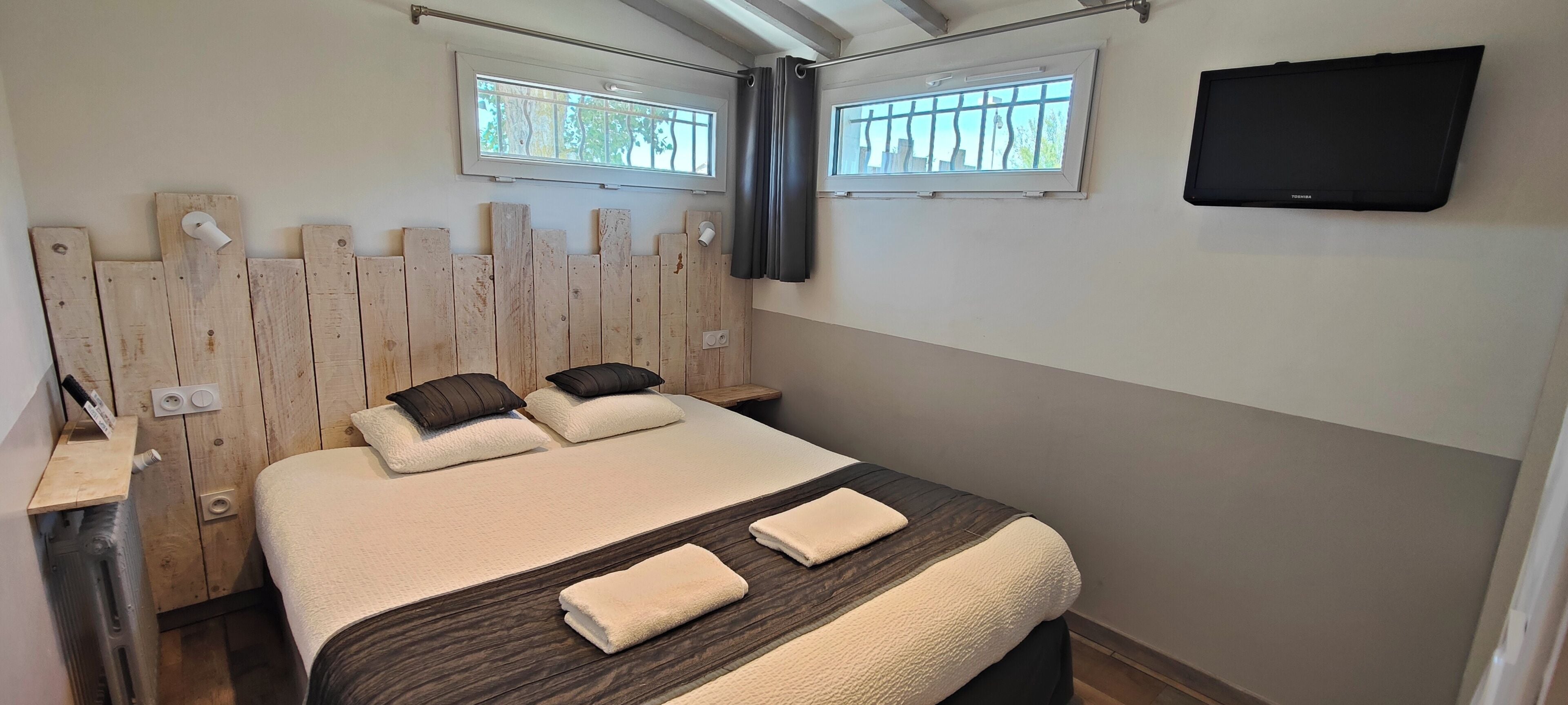 Un dormitorio con cabecera de madera, dos mesitas de noche, una cama con ropa de cama blanca y marrón oscuro, dos almohadas, un televisor de pantalla plana montado en la pared y una ventana con vistas a árboles.