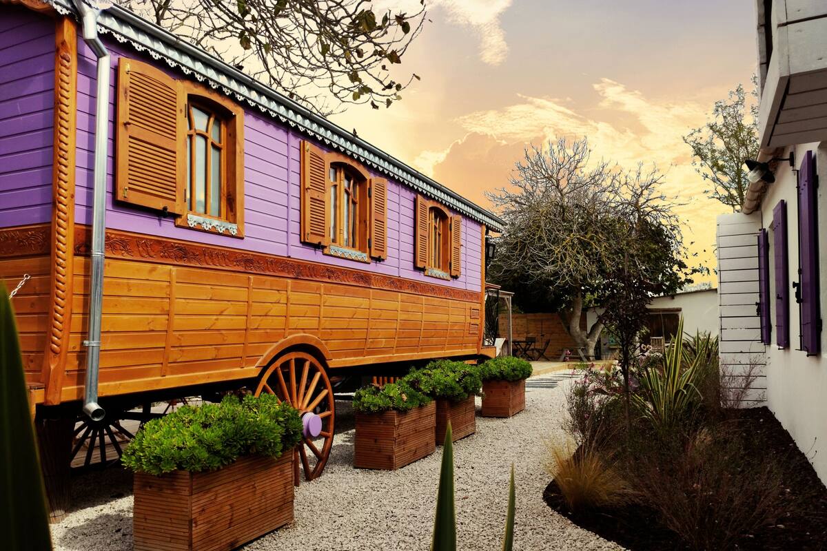 A purple and orange building with wooden shutters, a wheelbarrow, and planters in a gravel courtyard.