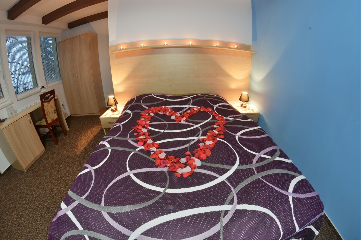 A bedroom with a bed, bedside lamps, a heart-shaped decoration on the bed, a wardrobe, and a window with blinds.