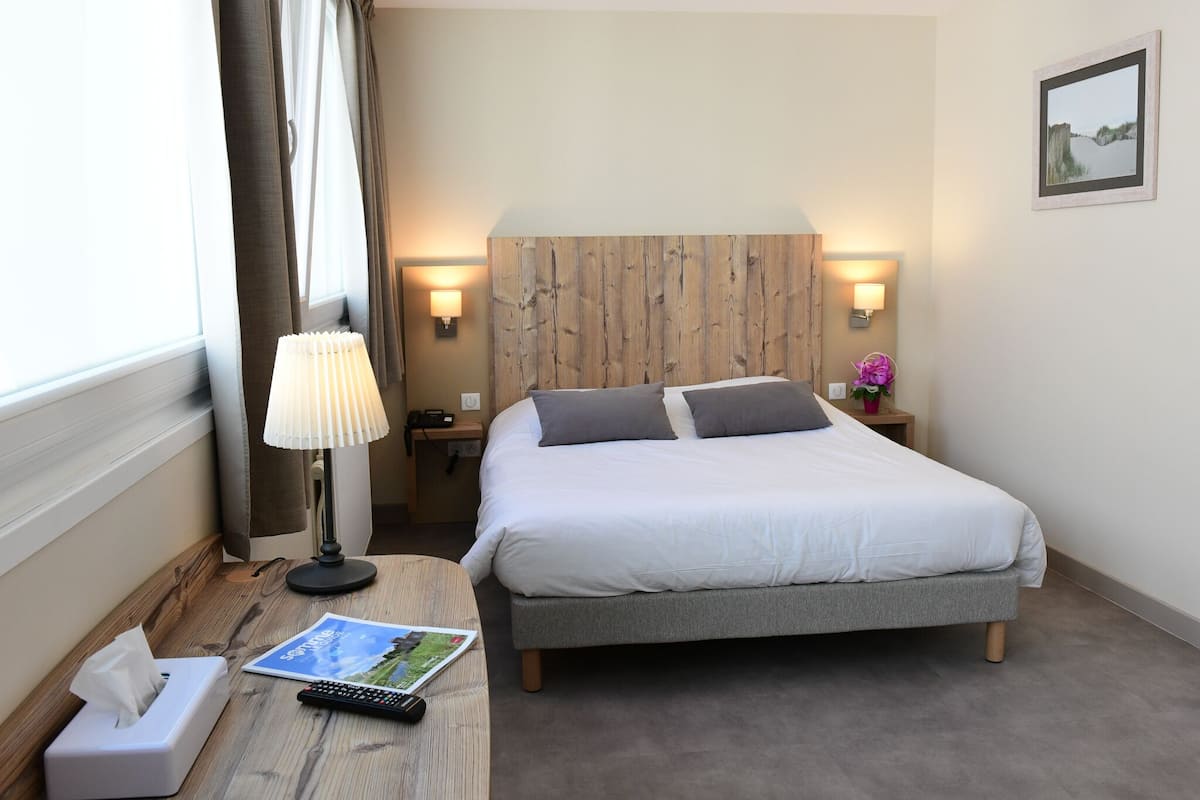 Una habitación de hotel ordenada con cama, lámpara de noche, mesita de noche y ventana con cortinas.