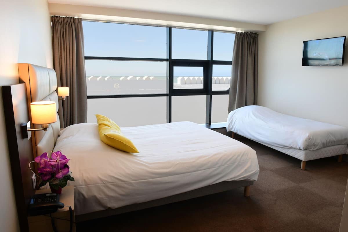 Habitación de hotel con un gran ventanal, una cama con ropa de cama blanca y un almohadón amarillo, una cama más pequeña, un televisor en la pared y un escritorio con lámpara y teléfono.