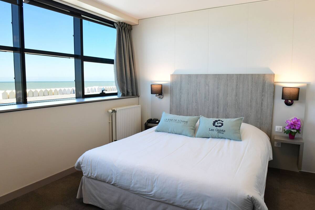 Un cuarto de hotel con una cama grande, dos lámparas de noche, una mesita de noche con un florero y vistas al mar a través de una ventana.