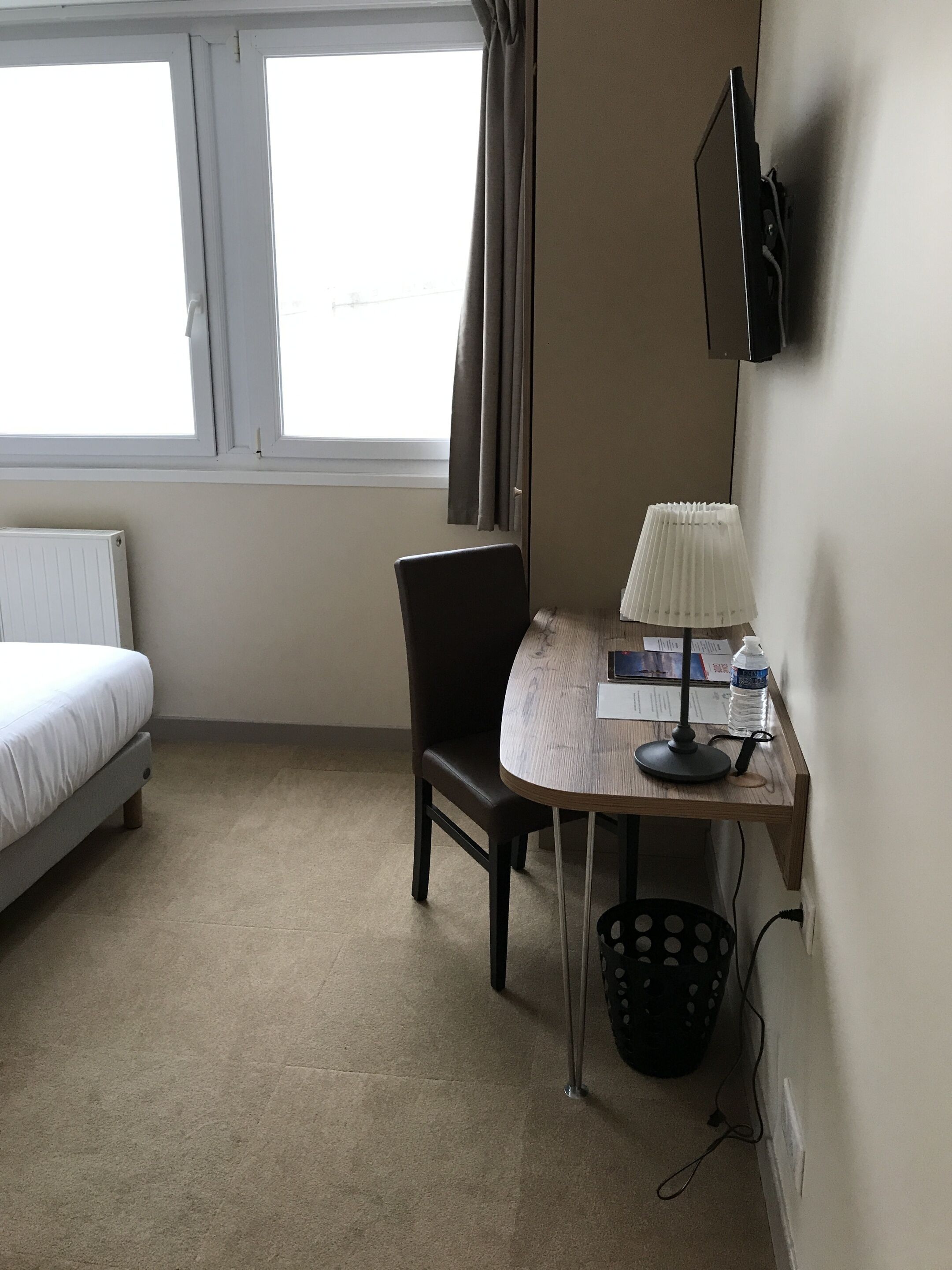 A hotel room with a bed, a desk with a lamp, a chair, a radiator, and a window with curtains.