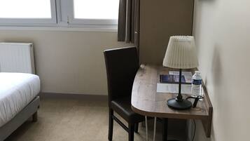 A hotel room with a bed, a desk with a lamp, a chair, a radiator, and a window with curtains.