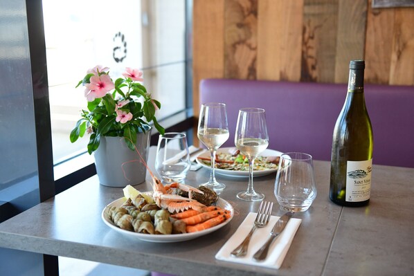 Una mesa servida con platos de comida, vasos y una botella de vino, con una planta en maceta y un banco morado al fondo.