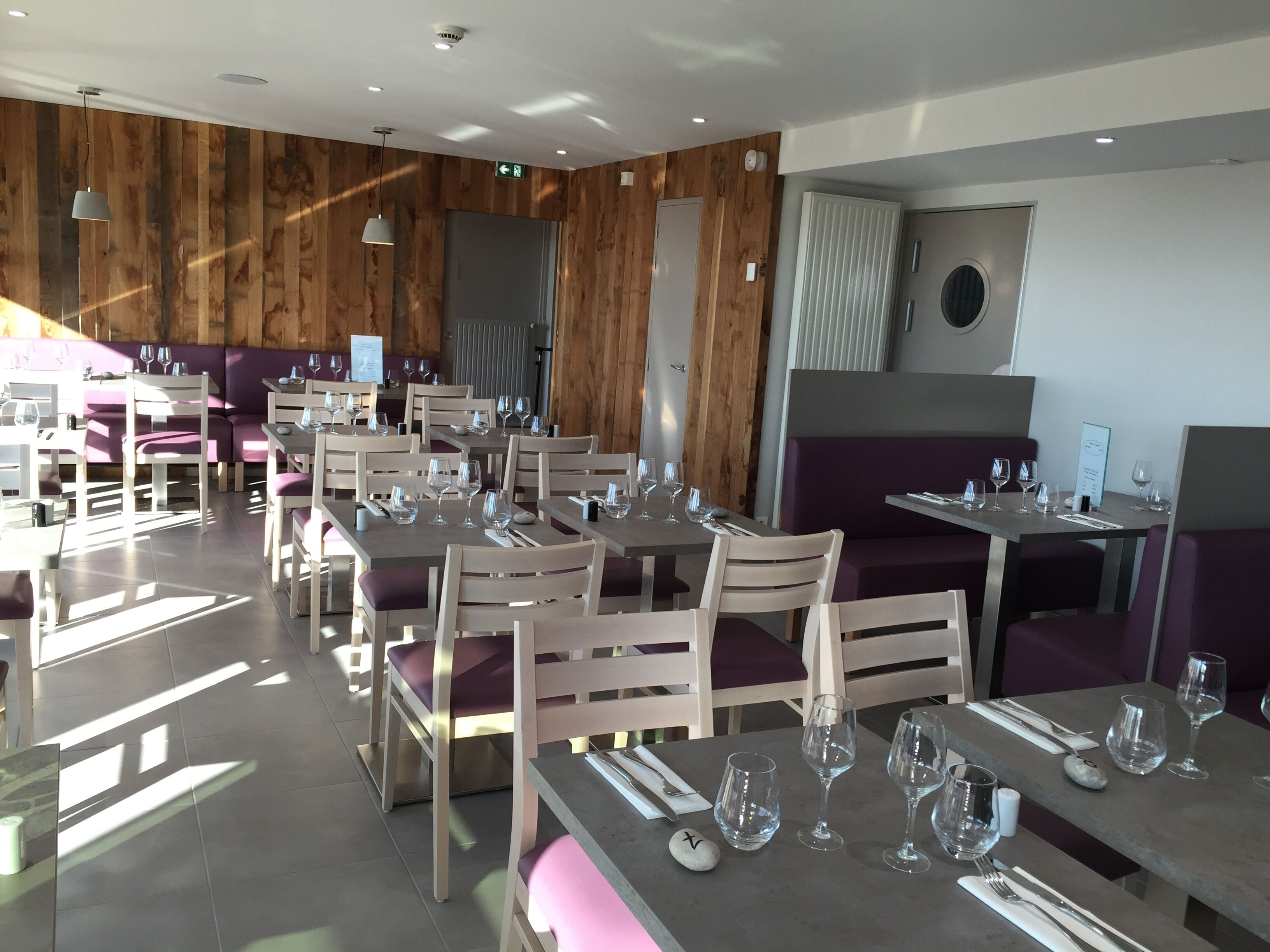 Un restaurante con paneles de madera, mesas dispuestas con copas de vino y cubiertos, y asientos con cojines morados.