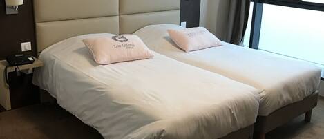 A double bed with two pillows, a nightstand with a lamp, a wall-mounted light, and a window with curtains.