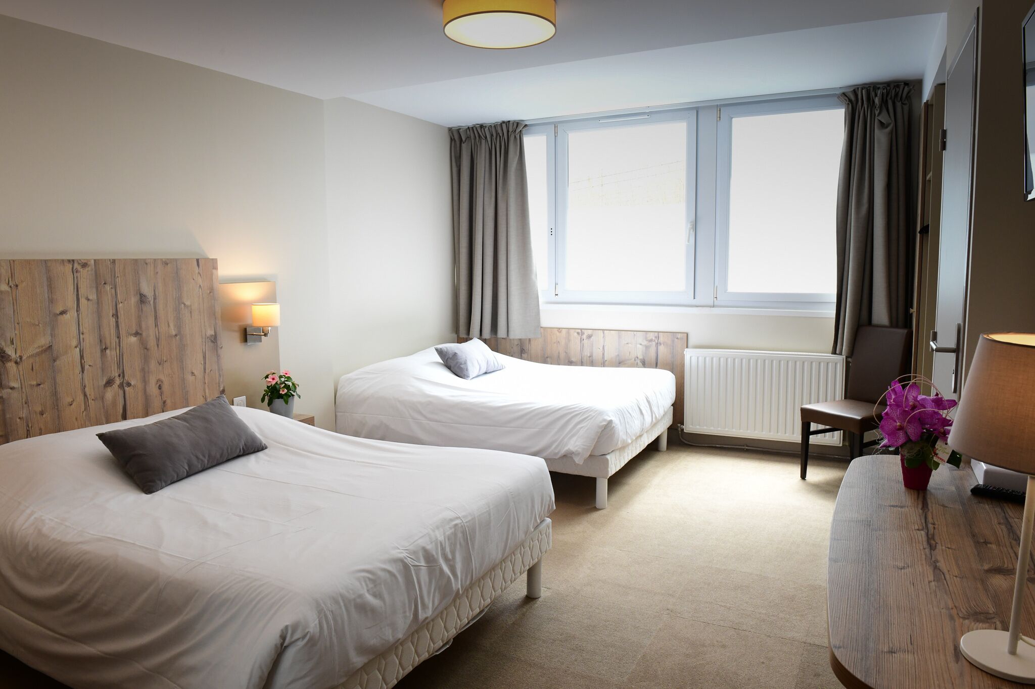 A hotel room with two beds, a wooden headboard, a window with curtains, a radiator, a desk with a lamp, and a chair.