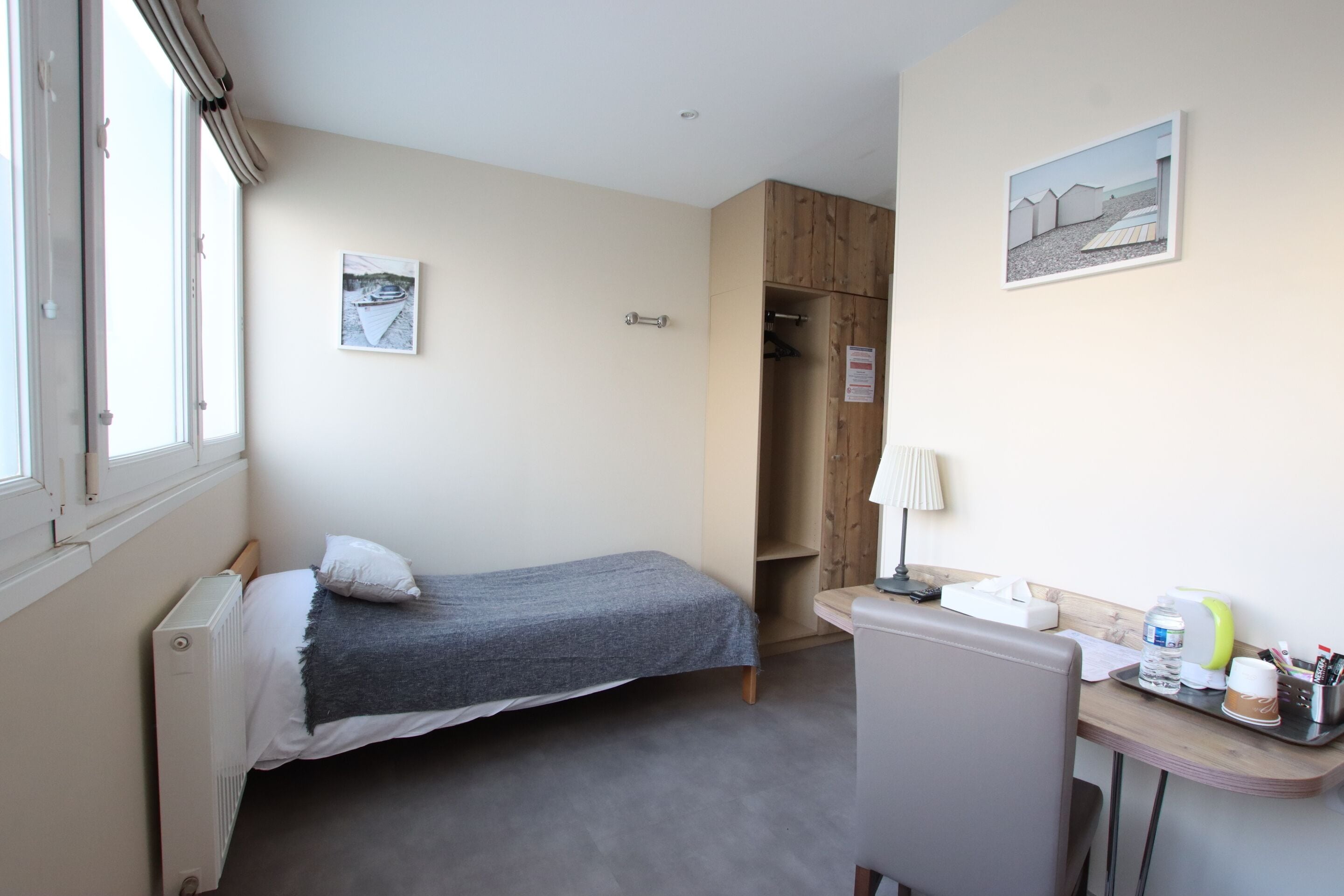 A small, neatly arranged bedroom with a single bed, a wooden wardrobe, a desk with a chair, and a window with blinds.