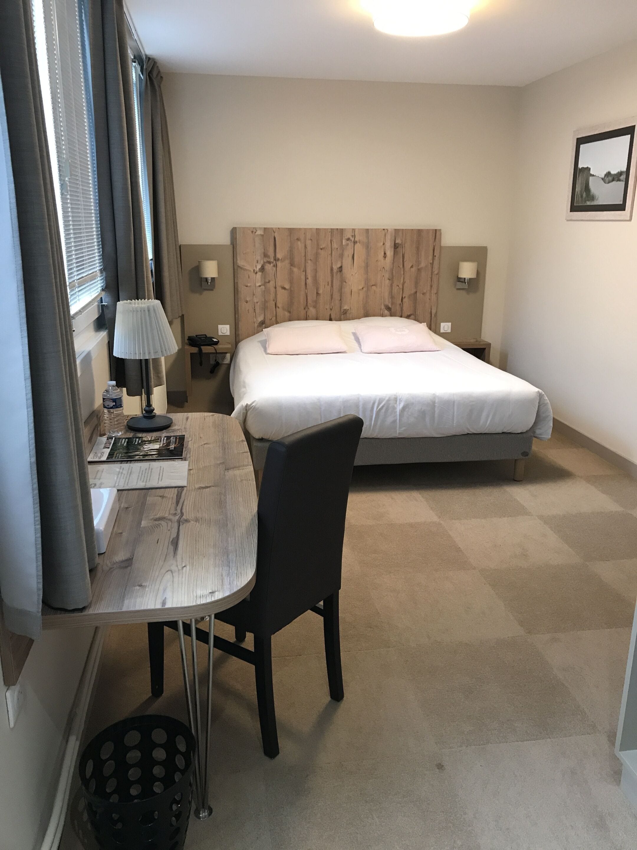 A hotel room with a bed, a desk with a chair, a lamp, and a small table.