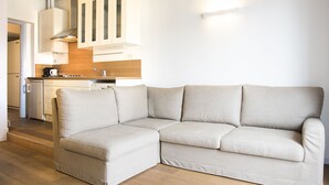 Uma sala de estar moderna com um sofá seccional bege, piso de madeira, e uma área de cozinha com armários brancos e forno embutido.