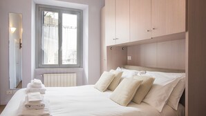 Uma cama bem arrumada com lençóis e almofadas brancas, uma janela com cortinas e um guarda-roupa de madeira.