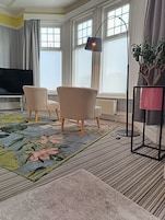Uma sala de estar com televisão de tela plana, duas poltronas, um abajur, uma planta em vaso e um tapete estampado.