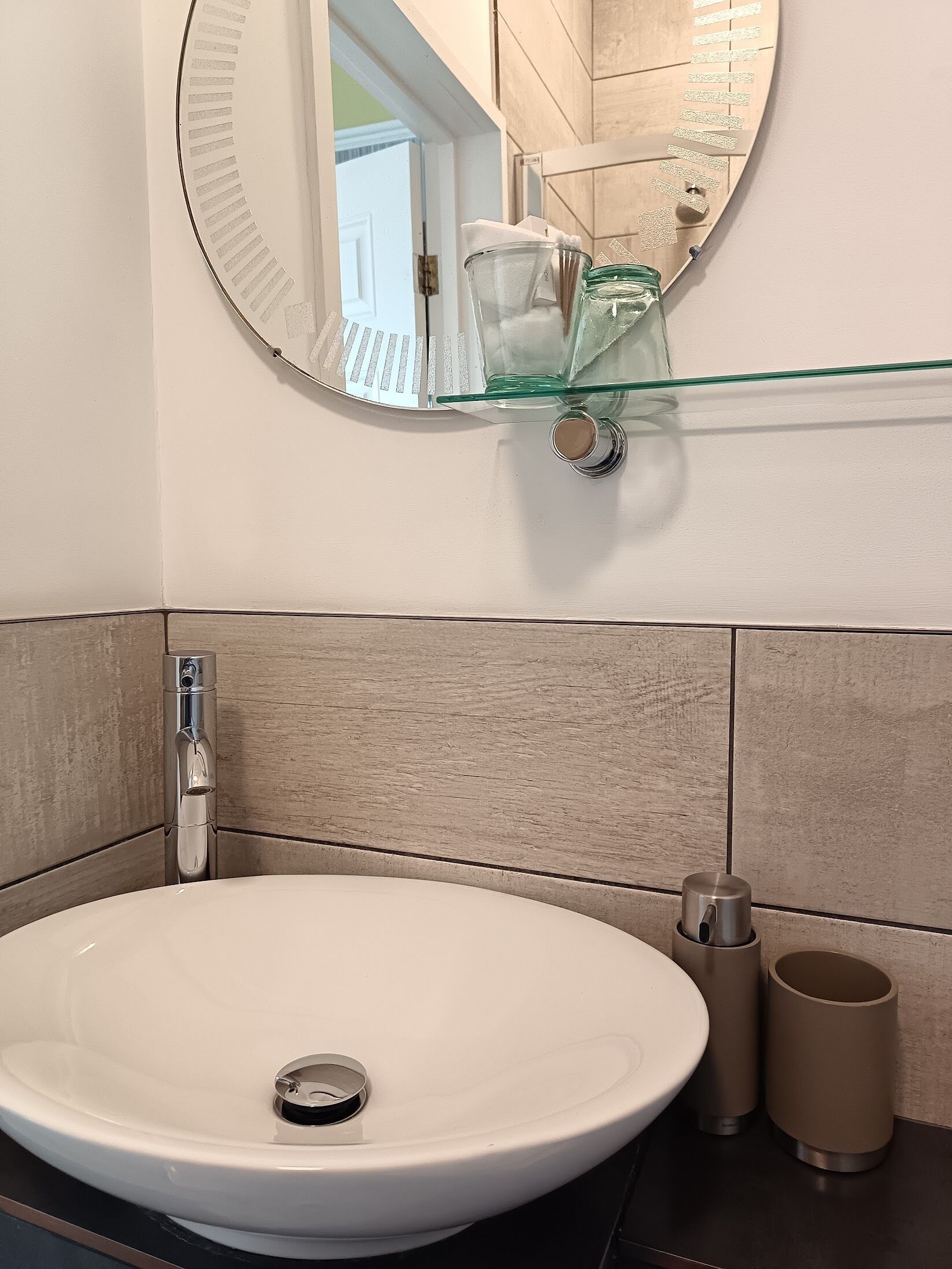 A bathroom with a round mirror, a glass shelf, a white sink, and a towel dispenser.