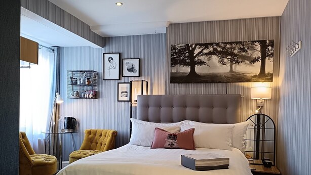A hotel room with a large bed, a bedside table with a lamp, a chair, and a wall-mounted shelf with framed pictures. - Regency Rooms (Littlehampton)