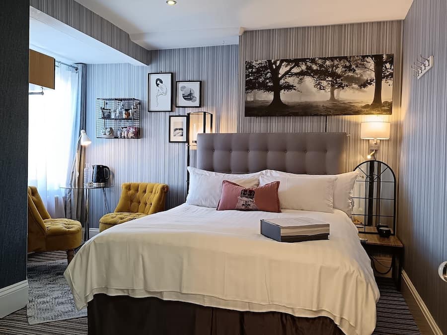 A hotel room with a large bed, a bedside table with a lamp, a chair, and a wall-mounted shelf with framed pictures.