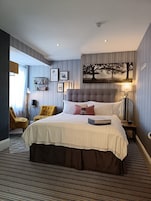 Quarto de hotel com uma cama grande, mesa de cabeceira com abajur, uma cadeira e prateleira na parede com quadros.