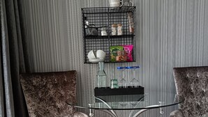 A room with two armchairs, a glass coffee table, a black metal shelf with various items, and a patterned wallpaper background. - Regency Rooms (Littlehampton)