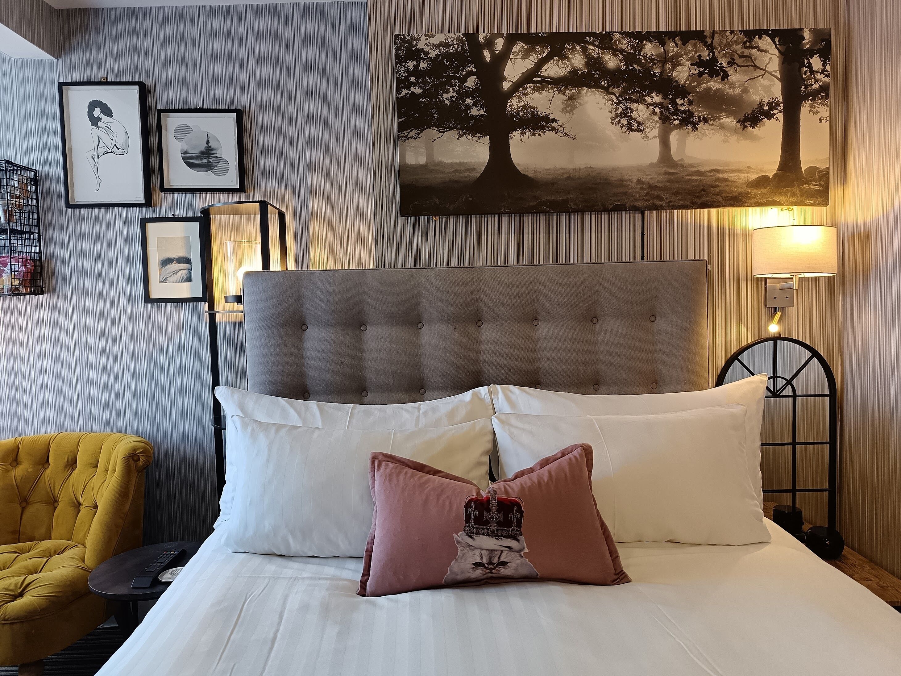 A hotel room with a tufted headboard, a bed with white linens and multiple pillows, a yellow armchair, a small round table with a remote control, and framed artwork on the wall.