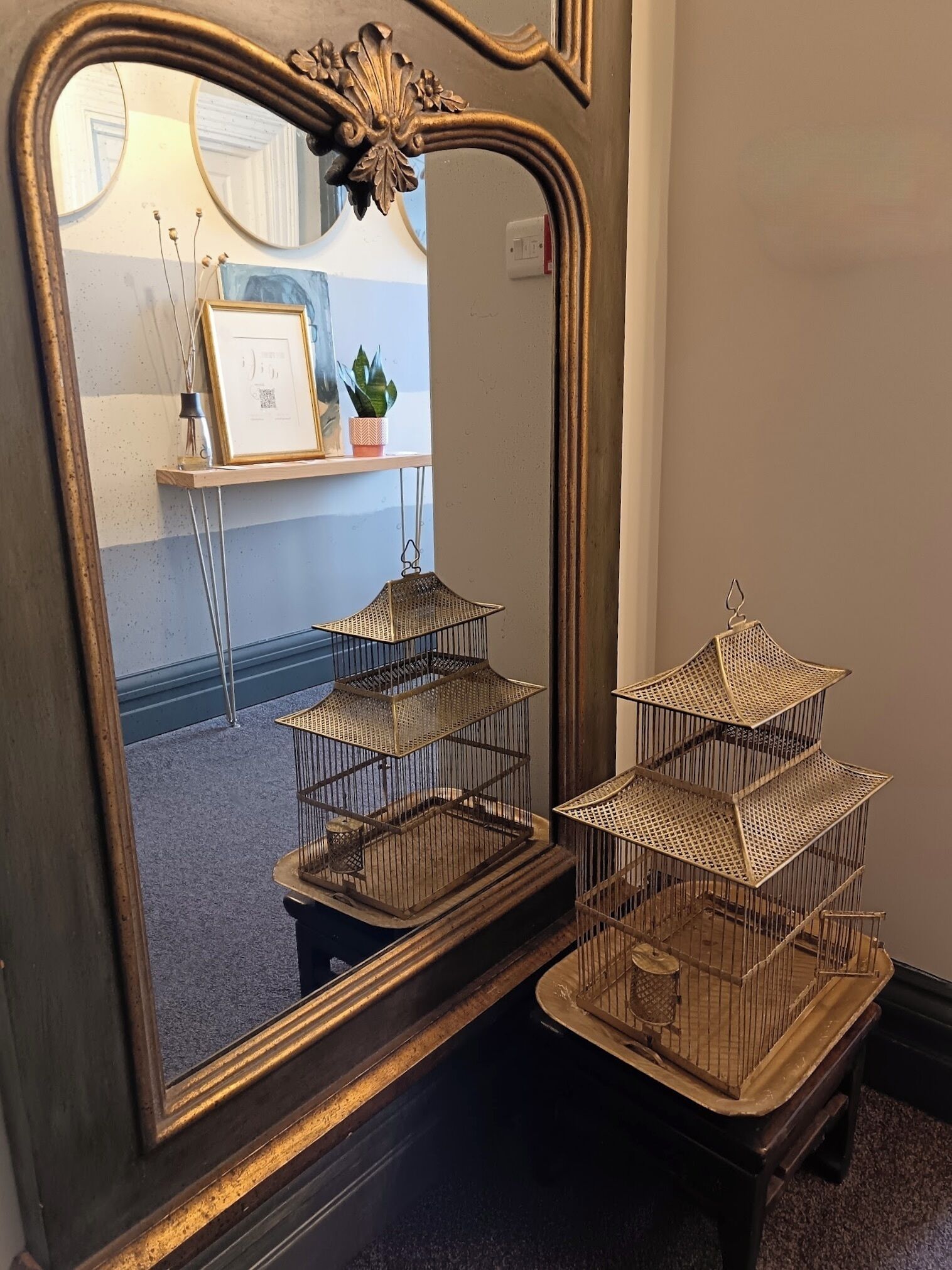 Un miroir aux détails dorés travaillés, qui reflète une pièce dotée d’un plancher en bois, d’un bureau avec une plante et d’un tableau encadré.