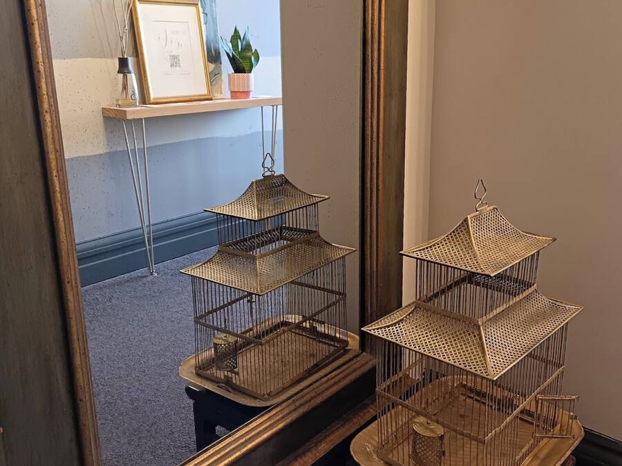 Un miroir aux détails dorés travaillés, qui reflète une pièce dotée d’un plancher en bois, d’un bureau avec une plante et d’un tableau encadré.
