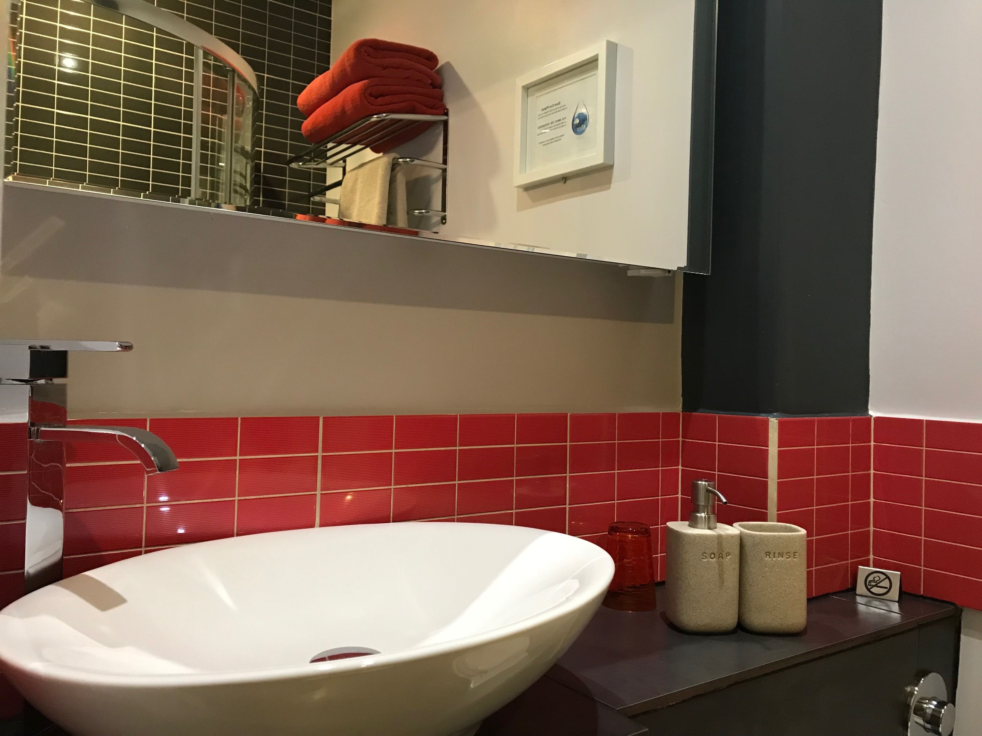 Un baño moderno con un lavamanos blanco, paredes de azulejos rojos, un toallero con toallas rojas y un dispensador de jabón.