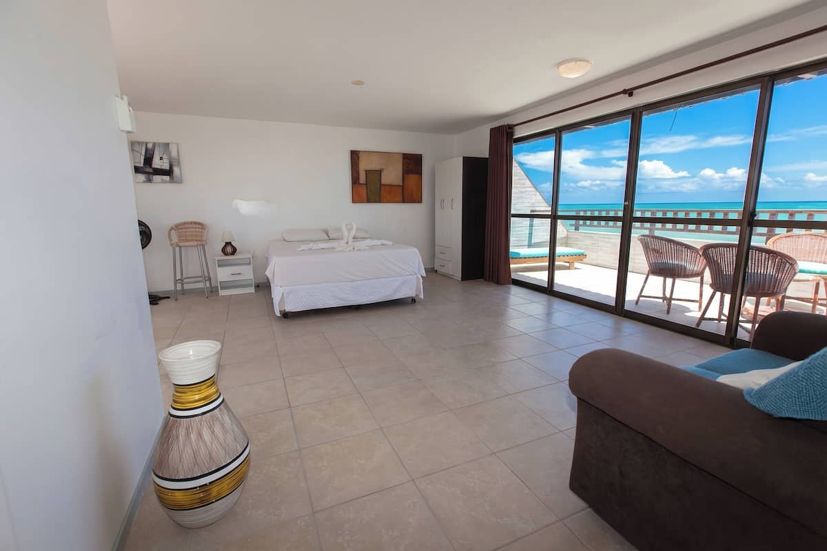 A hotel room with a bed, a large window with a view of the beach, a sitting area with a couch, and a decorative vase.