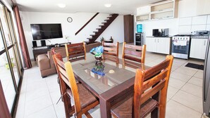 Una cocina moderna con zona de comedor, que incluye una mesa de vidrio, sillas de madera, horno empotrado y un televisor en la pared.
