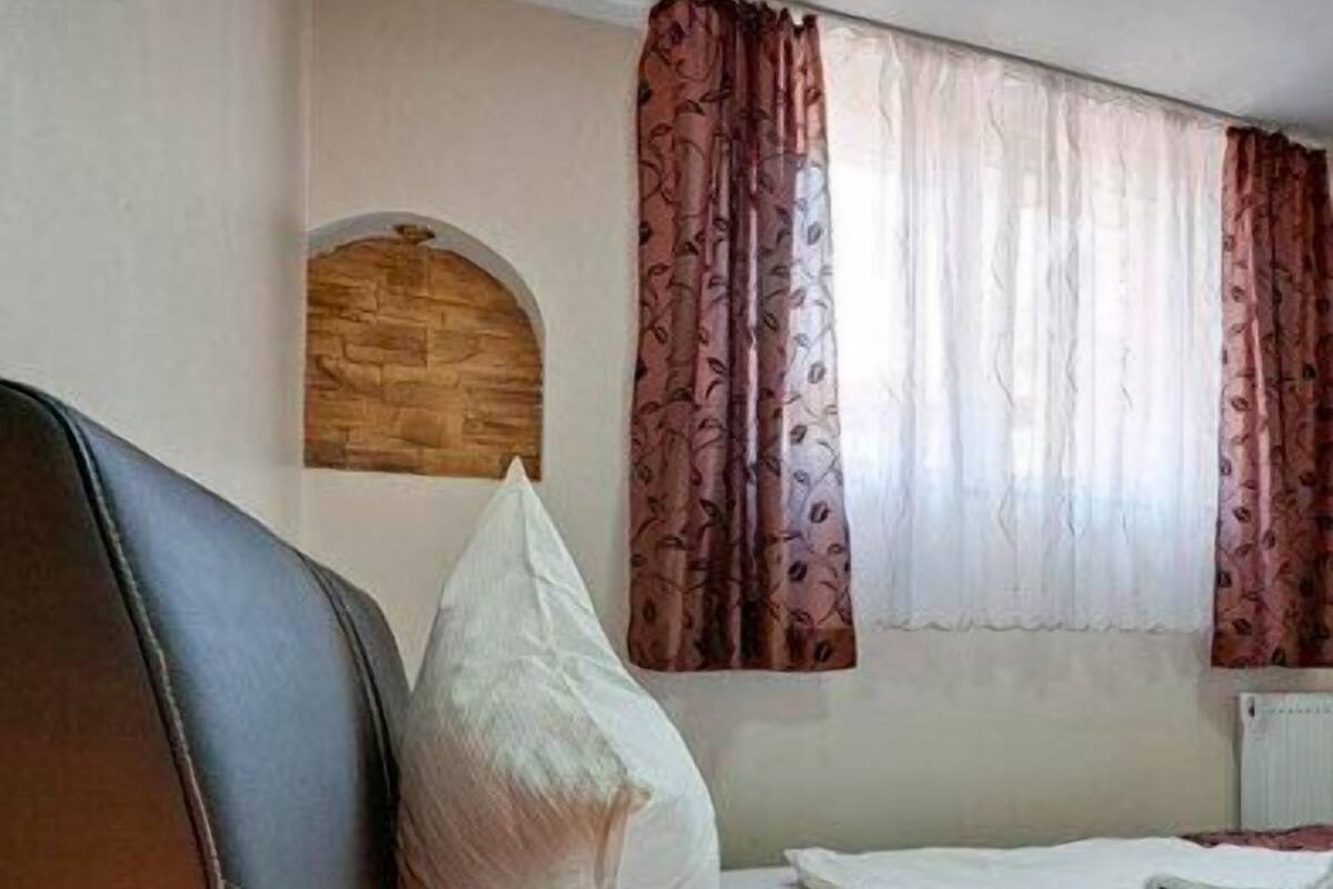 Een enkel bed met een witte matras, een wit kussen en een houten hoofdbord. Er is een raam met gordijnen met een patroon, een radiator eronder en een lamp op het nachtkastje.