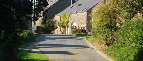 A narrow road leading to a stone building with a sloped roof, surrounded by greenery.