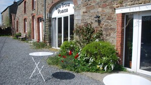 A stone building with a sign reading "Les Trois Voisins" and a gravel path with a small table and chairs. - B&B Les Trois Voisins (Gedinne)