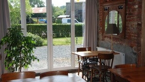 A dining area with a table and chairs, a potted plant, and a view of the outside through large sliding glass doors. - B&B Les Trois Voisins (Gedinne)