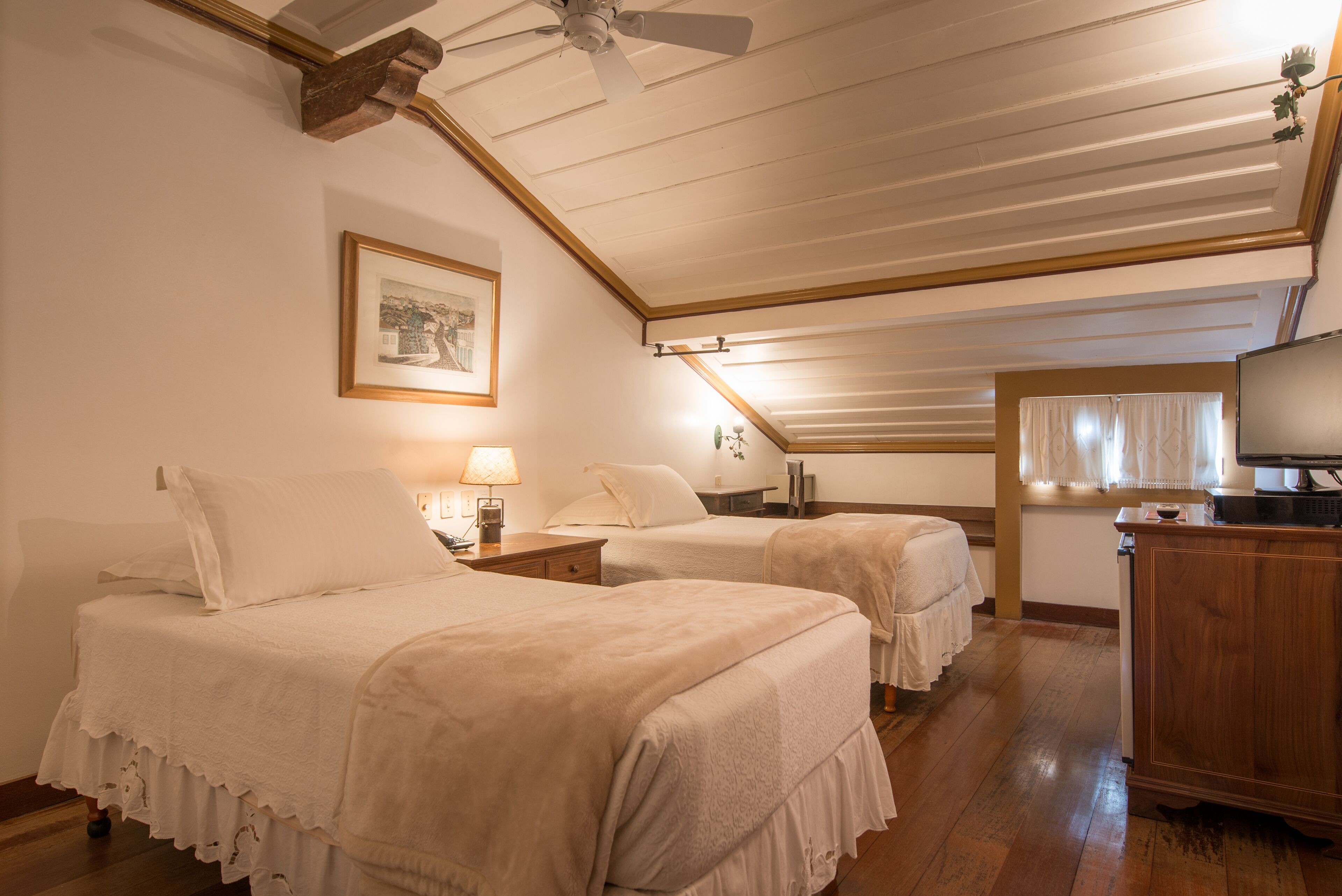 A room with two beds, a ceiling fan, a framed picture, a desk with a lamp, and a television.