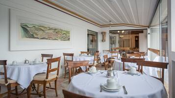 A dining area with round tables, wooden chairs, and white tablecloths. There is a large painting on the wall and a view of another room through a glass door.