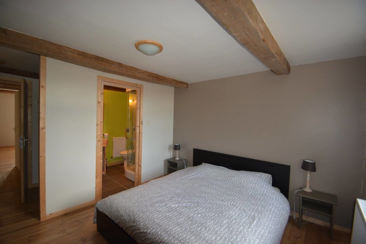 A bedroom with a bed, bedside tables, a nightstand, a wooden ceiling beam, and a door leading to a bathroom.