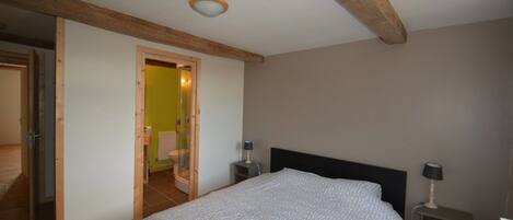 A bedroom with a bed, bedside tables, a nightstand, a wooden ceiling beam, and a door leading to a bathroom.