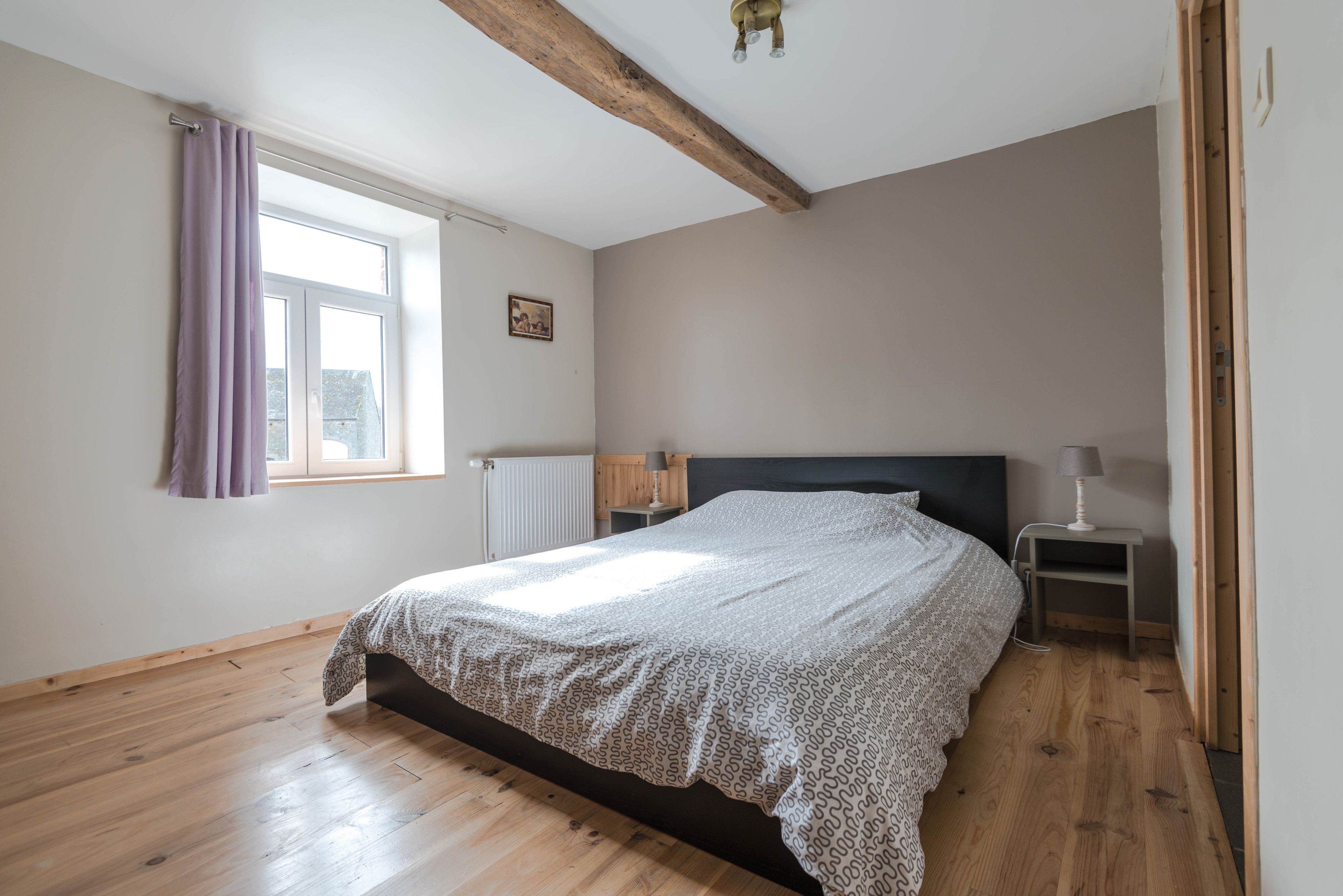 Un dormitorio con suelo de madera, una cama con colcha estampada, una ventana con cortinas moradas, una mesita de noche con lámpara y un radiador.