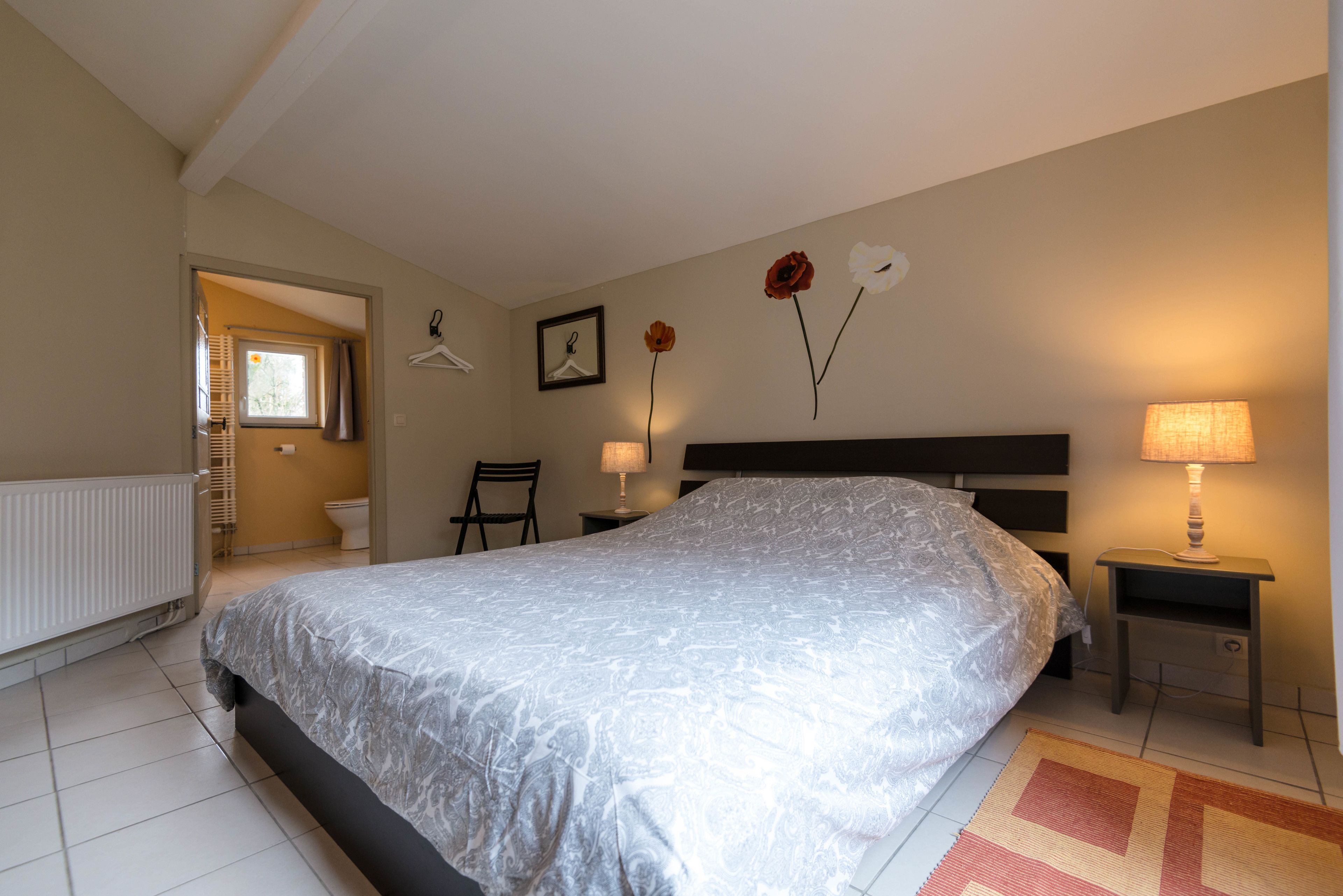 Een slaapkamer met een groot bed, een nachtkastje met een lamp, een stoel, een radiator en een raam met gordijnen.