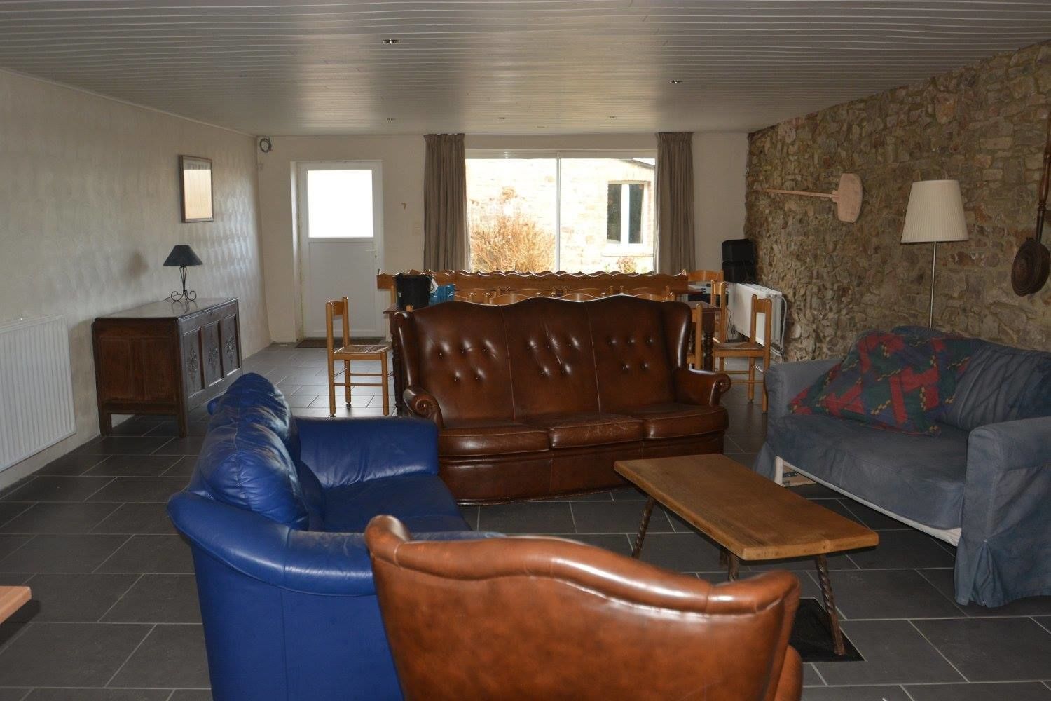 A living room with a brown leather sofa, a blue sofa, and a wooden coffee table. There is a stone wall and a window with curtains.