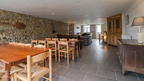 A spacious dining area with a stone wall, wooden tables and chairs, and a view into a living area with a sofa and wooden cabinets. - Gîte Les Trois Voisins (Gedinne)