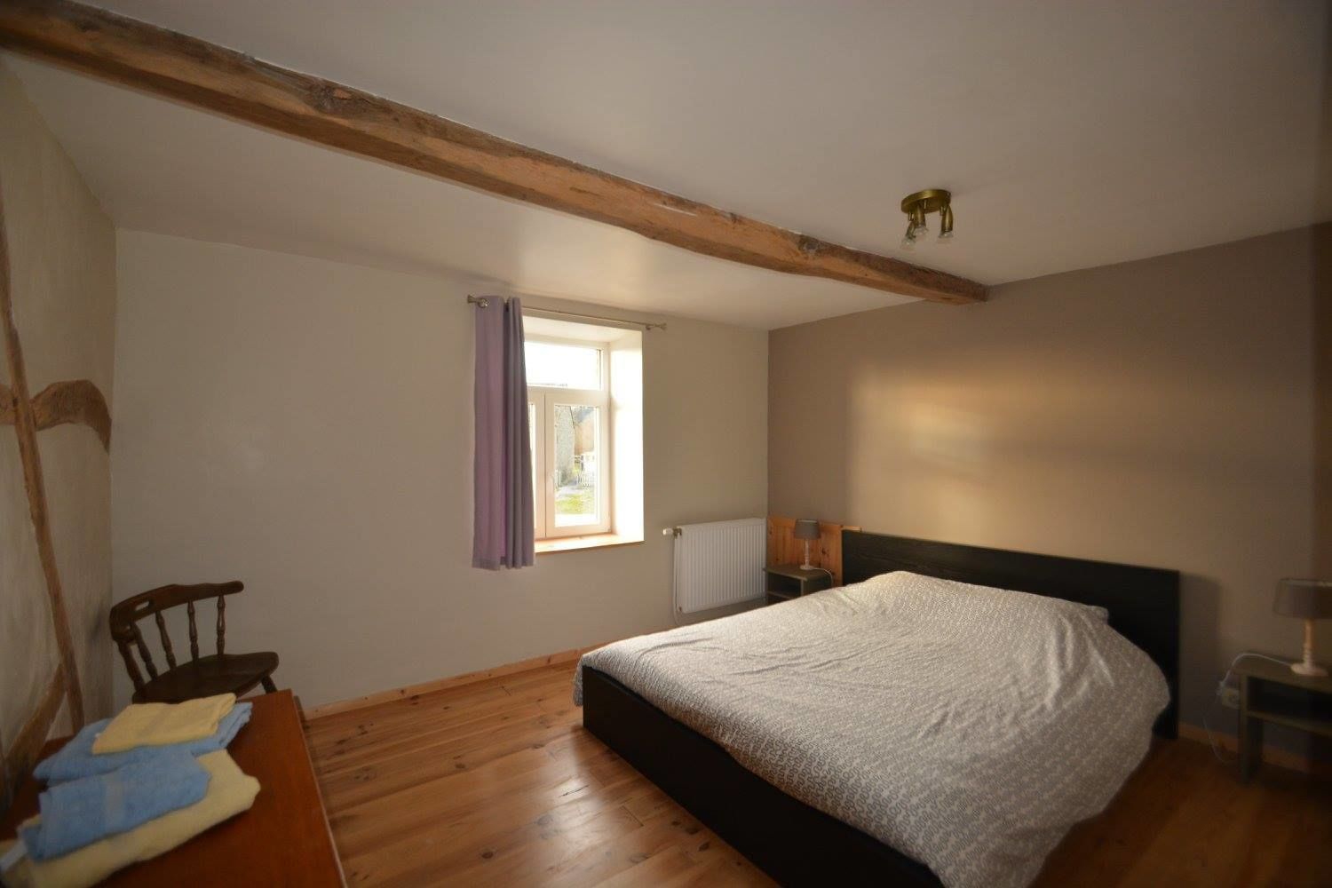 Un dormitorio con piso de madera, una cama con cabecera oscura, una ventana con cortinas y una silla.