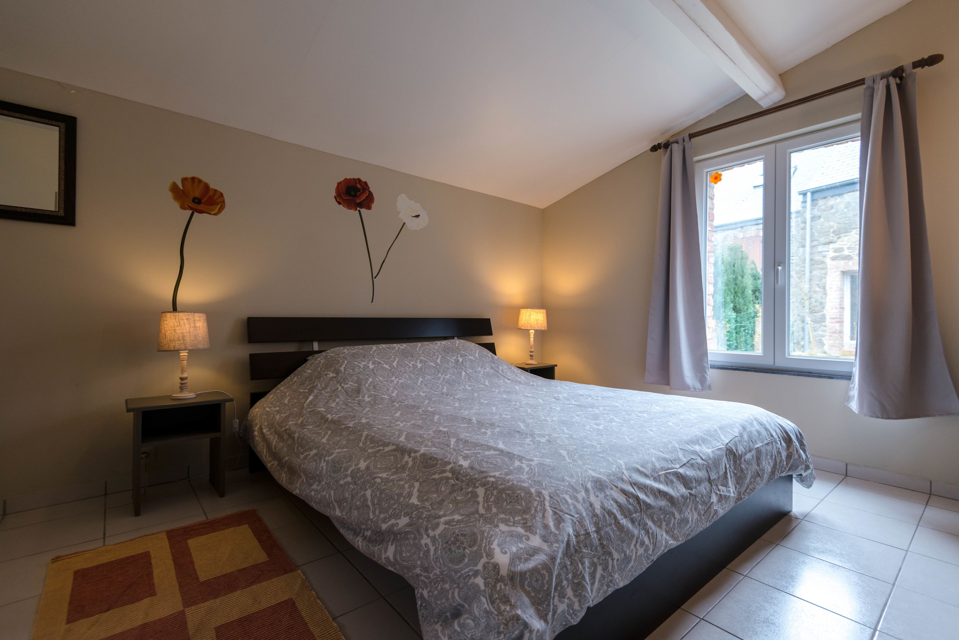 Un dormitorio con una cama grande, dos lámparas de noche, una mesita de noche, una alfombra estampada y un ventanal con cortinas.