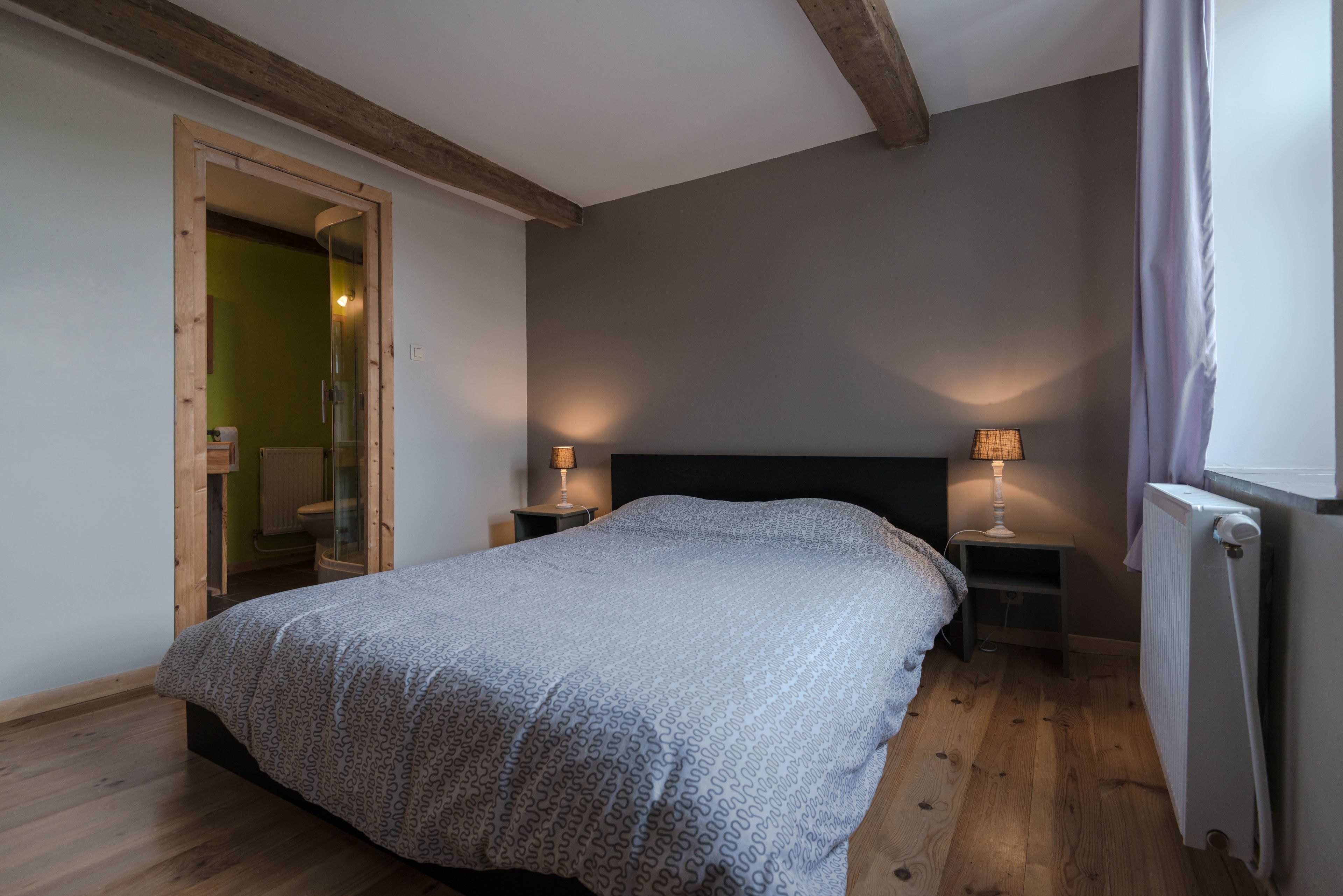 A bedroom with a bed, two bedside tables with lamps, a radiator, and a wooden door leading to another room.