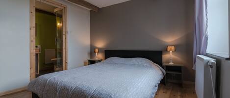 A bedroom with a bed, two bedside tables with lamps, a radiator, and a wooden door leading to another room.